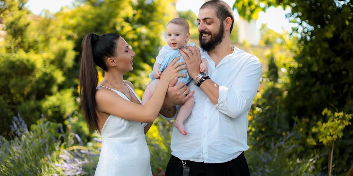 Mother and father joyfully hold their baby in a vibrant outdoor garden setting.