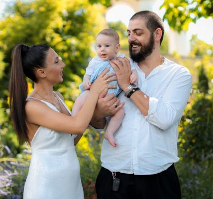 Mother and father joyfully hold their baby in a vibrant outdoor garden setting.