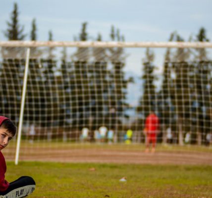 A boy sits cross-legged on a football field, wearing a hoodie and an orange vest.