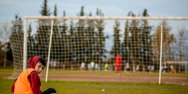 A boy sits cross-legged on a football field, wearing a hoodie and an orange vest.